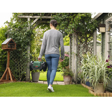 Frau geht mit einem Rasenmähroboter durch einen Garten mit Vogelhaus und Pflanzen