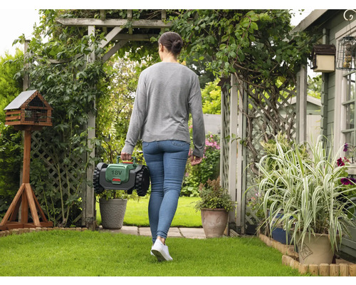 Frau geht mit einem Rasenmähroboter durch einen Garten mit Vogelhaus und Pflanzen