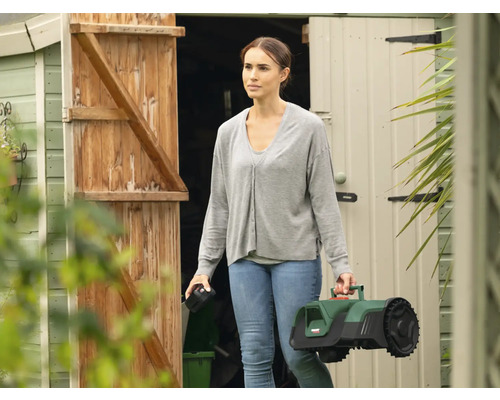 Frau trägt einen Rasenmähroboter vor einem Gartenschuppen.
