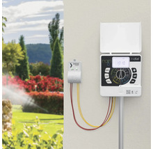 Rain i-dial Bewässerungscomputer und Smartizer Vision Modul an einer Wand montiert, im Hintergrund ein heller Garten mit aktivem Rasensprenger.