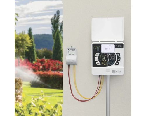 Rain i-dial Bewässerungscomputer und Smartizer Vision Modul an einer Wand montiert, im Hintergrund ein heller Garten mit aktivem Rasensprenger.