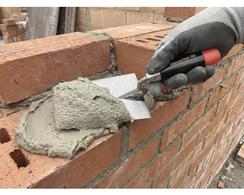 Maurerhand legt mit einer Maurerkelle Mörtel auf eine Ziegelsteinmauer.