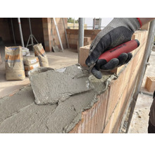 Maurer arbeitet mit einer Maurerkelle an einer Ziegelwand, im Hintergrund Zementsäcke.