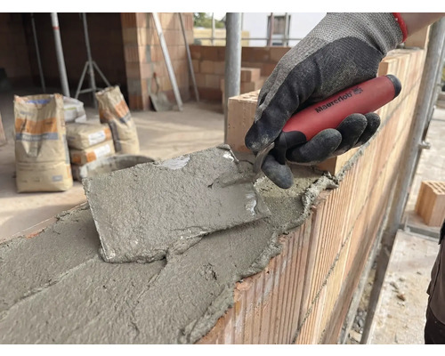 Maurer arbeitet mit einer Maurerkelle an einer Ziegelwand, im Hintergrund Zementsäcke.