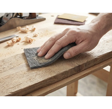 Hand bei der Bearbeitung eines Holzbretts mit Schleifvlies auf einer hellen Werkbank mit Hobel und Holzspänen.