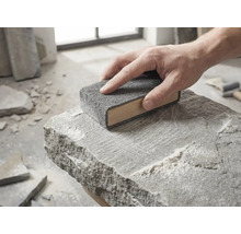 Eine Hand führt einen hölzernen Schleifklotz mit grauem Schleifvlies über die Oberfläche eines rauen Natursteins.