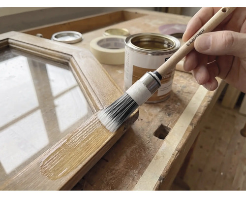 Anwendung einer Holzlasur auf einem Holzrahmen mit einem Rautner Rundpinsel auf einer hellen Werkbank mit Farbdose und Klebeband.