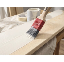 Ein Pinsel mit Holzstiel trägt weiße Farbe auf ein Holzbrett auf, im Hintergrund sind ein Farbeimer und Klebeband zu sehen.