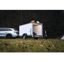 Weißer Humbaur Kofferanhänger an einem Auto auf einer Waldwiese, mit geöffneter Hecktür, Innenbeleuchtung und Campingausrüstung.