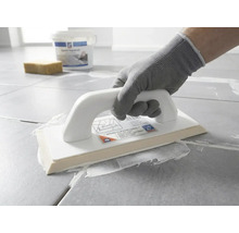 Eine Person mit Schutzhandschuh trägt mit einem Epoxid-Fugbrett weiße Fugenmasse auf graue Bodenfliesen auf.