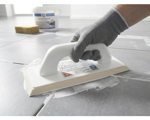 Eine Person mit Schutzhandschuh trägt mit einem Epoxid-Fugbrett weiße Fugenmasse auf graue Bodenfliesen auf.