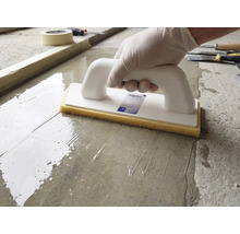 Eine Person nutzt ein Schwammbrett mit weißem Griff und gelbem Schwamm auf einer feuchten Zementoberfläche.