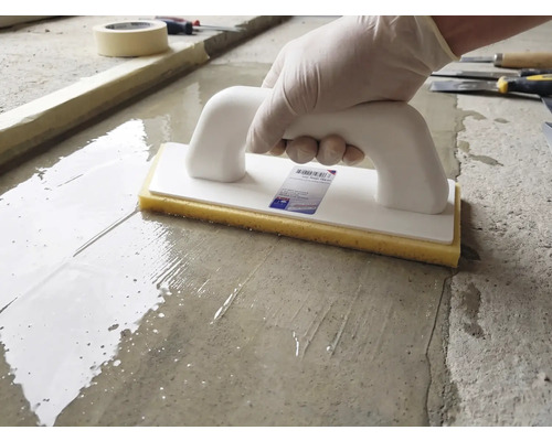 Eine Person nutzt ein Schwammbrett mit weißem Griff und gelbem Schwamm auf einer feuchten Zementoberfläche.