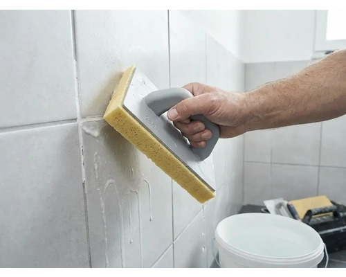 Eine Person reinigt frisch verfugte Wandfliesen mit einem Schwammbrett und Wasser.