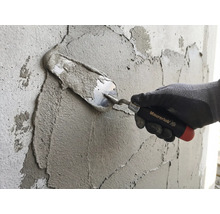 Eine Hand mit Arbeitshandschuh trägt mit einer Maurerlob Maurerkelle Mörtel auf eine Wand auf.