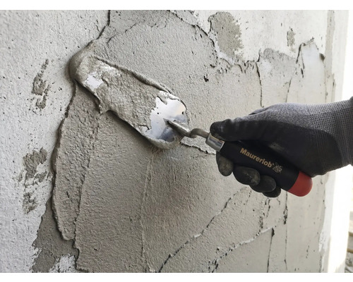 Eine Hand mit Arbeitshandschuh trägt mit einer Maurerlob Maurerkelle Mörtel auf eine Wand auf.