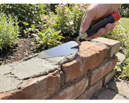 Hand mit Maurerlob Maurerkelle trägt Mörtel auf eine Ziegelmauer im Garten auf.