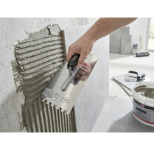 Eine Hand trägt mit einer Glättkelle mit Zahnung Fliesenkleber auf eine Wand auf.