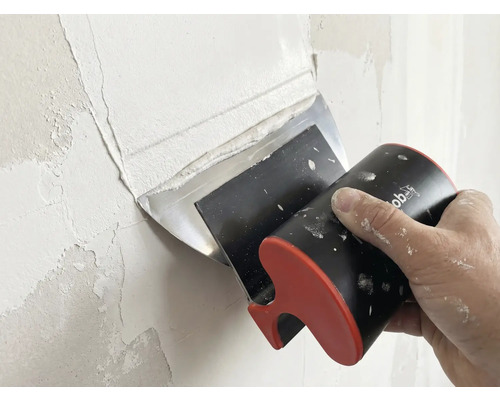 Hand trägt mit einem Flächenspachtel Spachtelmasse auf eine Wand auf.