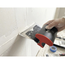 Hand führt einen Breitspachtel zum Auftragen und Glätten von weißer Spachtelmasse über einer Wandfuge.