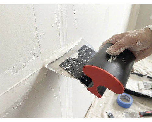 Hand führt einen Breitspachtel zum Auftragen und Glätten von weißer Spachtelmasse über einer Wandfuge.