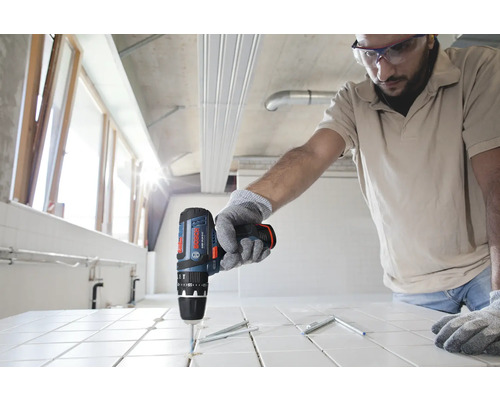 Person verwendet einen Bosch Akku-Schlagbohrschrauber zum Bohren in weiße Fliesen auf einer Baustelle.
