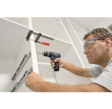 Ein Mann mit Schutzbrille montiert Schubladenschienen in einem weißen Schrank mit einem Bosch Akku-Bohrschrauber und einer Schraubzwinge.