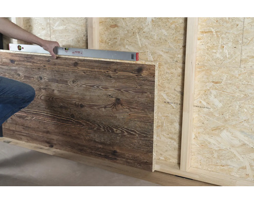Eine Person richtet ein Wandpaneel in Holzoptik mit einer Wasserwaage an einer Wand aus Grobspanplatten aus.