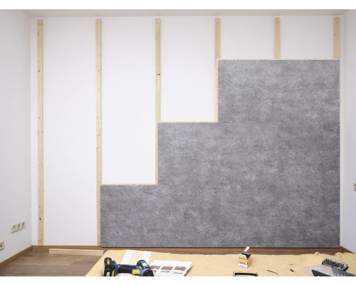 Montage grauer Wandpaneele in Betonoptik auf einer Holzlatten-Unterkonstruktion an einer weißen Wand im Innenraum.