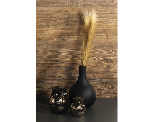 Schwarze Vase mit Pampasgras neben zwei Eulenfiguren vor einer Holzwand.