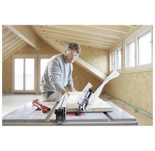 Ein Mann mit Schutzbrille schneidet ein Holzbrett mit einer Tischkreissäge in einem hellen Raum mit Holzbalken und OSB-Wänden.