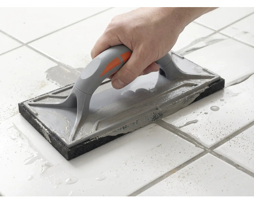 Eine Hand führt ein Fugenbrett über weiße Bodenfliesen, um graue Fugenmasse zu verteilen.