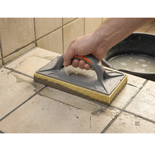 Ein Schwammbrett wird zum Reinigen von frisch verfugten Fliesen verwendet. Eine Hand führt das Brett über die nassen Fliesen.
