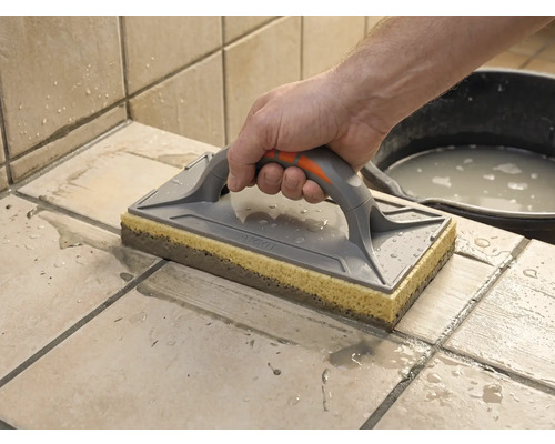 Ein Schwammbrett wird zum Reinigen von frisch verfugten Fliesen verwendet. Eine Hand führt das Brett über die nassen Fliesen.