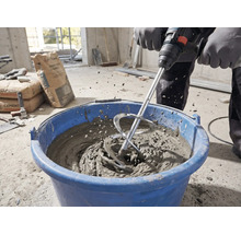 Person mischt Mörtel mit einem Rührkorb und einem Rührwerk in einem blauen Eimer auf einer Baustelle.