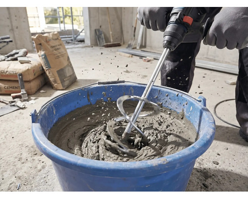 Person mischt Mörtel mit einem Rührkorb und einem Rührwerk in einem blauen Eimer auf einer Baustelle.