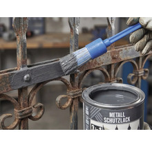 Metallschutzlack wird mit einem Rundpinsel auf einen rostigen Metallzaun aufgetragen.