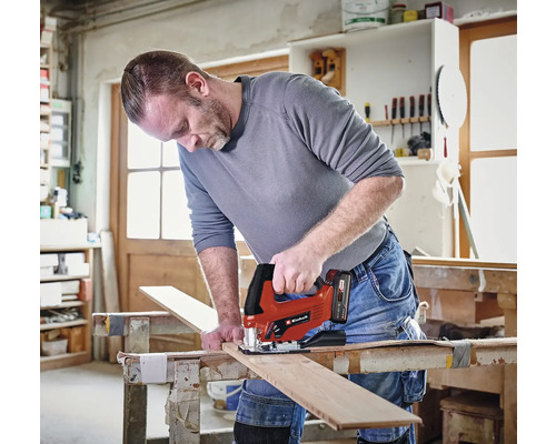 Ein Handwerker schneidet in einer hellen Werkstatt mit einer batteriebetriebenen Einhell Stichsäge ein Holzbrett auf einer Werkbank. Einhell Logo.