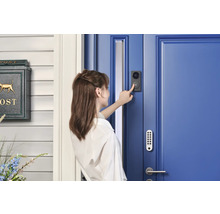 Eine Frau betätigt eine schwarze Video-Türsprechanlage an einer blauen Haustür mit Tastaturschloss neben einem grünen Briefkasten.