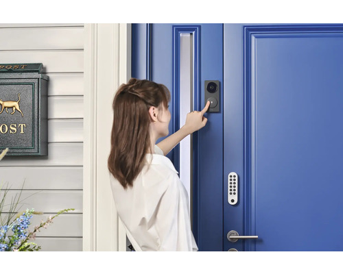 Eine Frau betätigt eine schwarze Video-Türsprechanlage an einer blauen Haustür mit Tastaturschloss neben einem grünen Briefkasten.