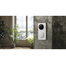 Weiße Video-Türklingel mit schwarzer Kamera und runder Klingeltaste, montiert an einer strukturierten Wand.