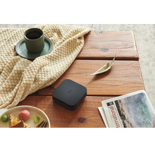 Aqara Smart Home Hub M2 auf einem Holztisch neben einer Tasse Kaffee auf einer Strickdecke, einem Teller mit Obst und einer Zeitschrift.