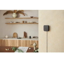 Schwarzer Smart Home Hub an einer hellen Wand in einer modern eingerichteten, hellen Wohnumgebung mit Holzregalen und Dekoration.