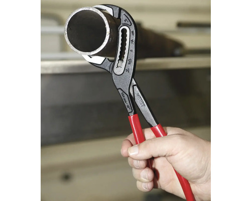 Eine Hand hält eine Virax Wasserpumpenzange mit roten Griffen, die ein Metallrohr umschließt.