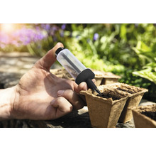 Eine Hand nutzt eine Sämaschine zum Aussäen von Samen in biologisch abbaubare Anzuchttöpfe in einem hellen Garten.