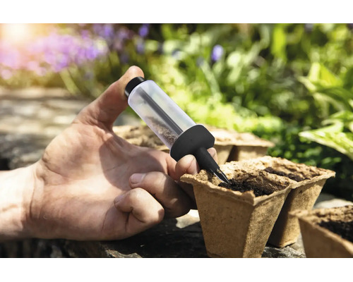 Eine Hand nutzt eine Sämaschine zum Aussäen von Samen in biologisch abbaubare Anzuchttöpfe in einem hellen Garten.
