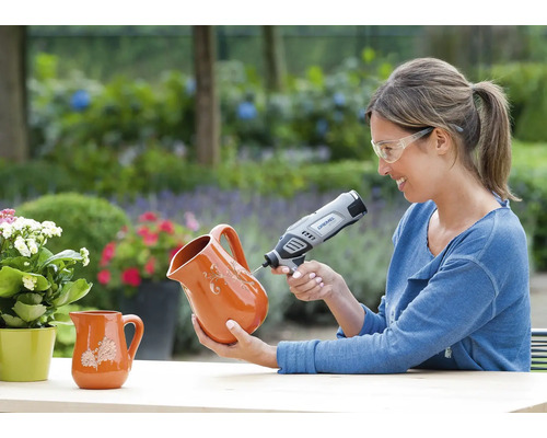 Frau mit Schutzbrille graviert einen Krug aus Terrakotta mit einem Dremel Multifunktionswerkzeug in einem hellen Garten.