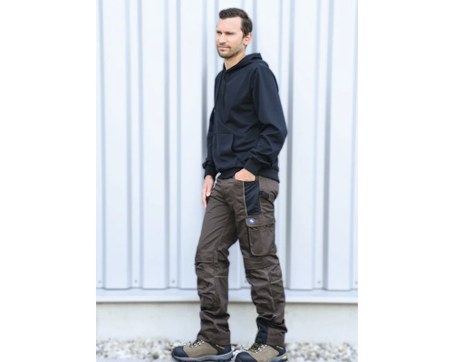 Mann in brauner Arbeitshose mit verstärkten Knien und Taschen, kombiniert mit schwarzem Kapuzenpullover und Arbeitsschuhen.