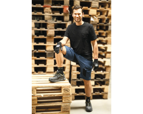 Mann in blauen Arbeitsshorts, schwarzem T-Shirt, Arbeitshandschuhen und Sicherheitsschuhen vor gestapelten Holzpaletten.