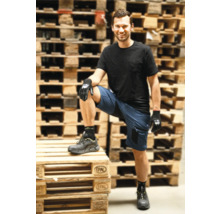 Mann in blauen Arbeitsshorts, schwarzem T-Shirt, Arbeitshandschuhen und Sicherheitsschuhen vor gestapelten Holzpaletten.